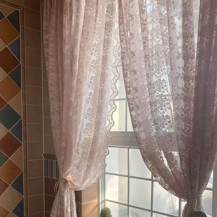 Voilage en dentelle rose clair à motifs floraux délicats, attaché partiellement pour laisser entrer la lumière naturelle dans une pièce carrelée chaleureuse.