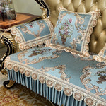 Un canapé d'inspiration baroque, avec des motifs floraux et des ornements ornés sur un fond bleu clair, complété par des coussins assortis et une frange délicate.