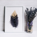 Une plume ornementée, dorée et décorative, posée dans un cadre blanc, avec un bouquet de lavande violette dans un vase en verre à côté.