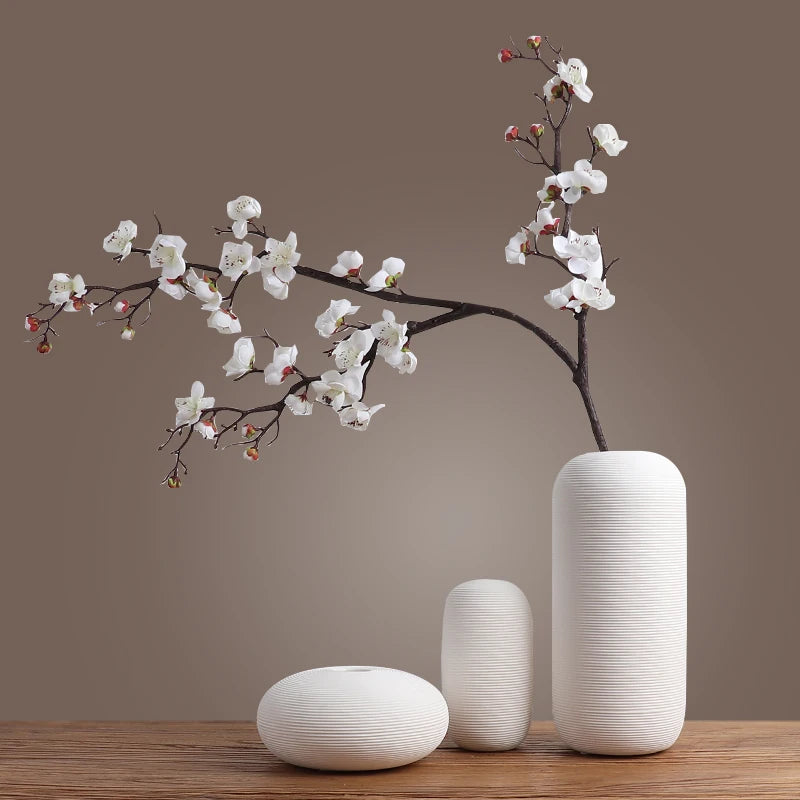 Une branche de fleurs de cerisier épanouis, avec de délicates fleurs blanches et roses, se dresse gracieusement dans de belles jarres cylindriques blanches sur une table en bois sombre.