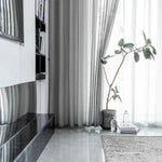 Intérieur moderne et épuré avec des rideaux gris élégants. Plante en pot ajoutant une touche naturelle à la pièce. Le sol brillant et le tapis doux créent une ambiance sophistiquée. Étagères remplies de livres apportant une dimension culturelle à l'espace de vie.