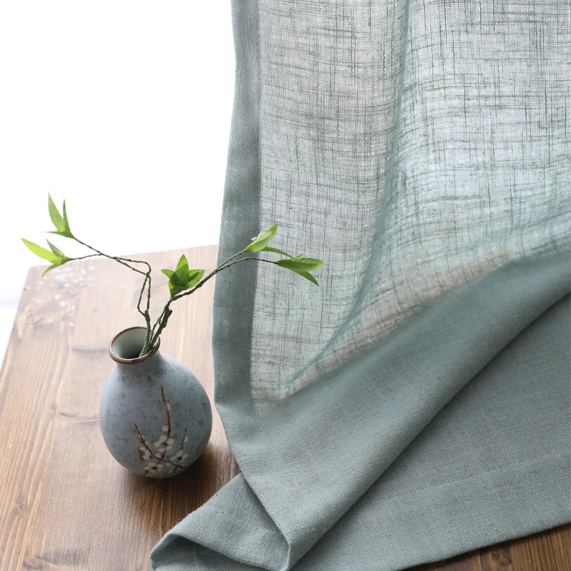 Voilage en tulle gris transparent tombant délicatement sur une table en bois avec un petit vase bleu et des branches décoratives.