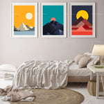 Trois affiches géométriques minimalistes représentant des paysages montagneux aux couleurs vives, suspendues au-dessus d'un lit confortable dans une chambre lumineuse.