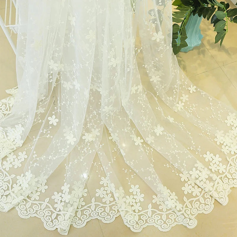 Détail du voilage blanc brodé de fleurs, étendu au sol avec ourlet festonné élégant, capturant la lumière naturelle sur carrelage clair.