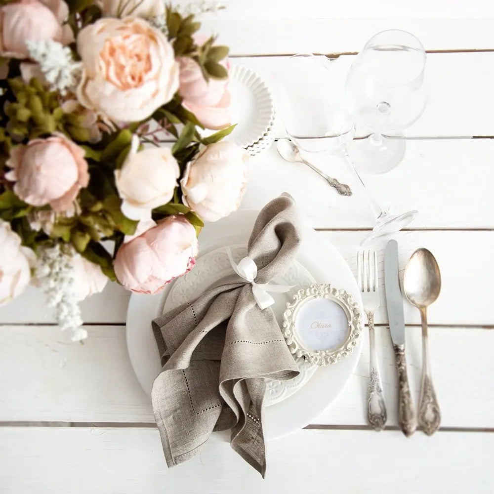 Sur une table blanche se trouve un arrangement floral de pivoines roses délicates, accompagné d'un couvert élégant avec serviette nouée et cadre photo.