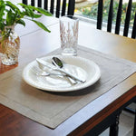 Un plan de table élégant avec une assiette en céramique blanche, des couverts argentés et un verre de cristal décoré, le tout placé sur un dessous de table gris neutre.