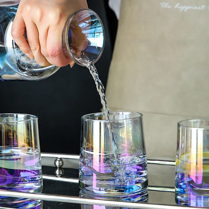 L'image montre un barman versant un liquide coloré dans des verres transparents, créant un effet visuel intéressant avec les reflets et les couleurs.