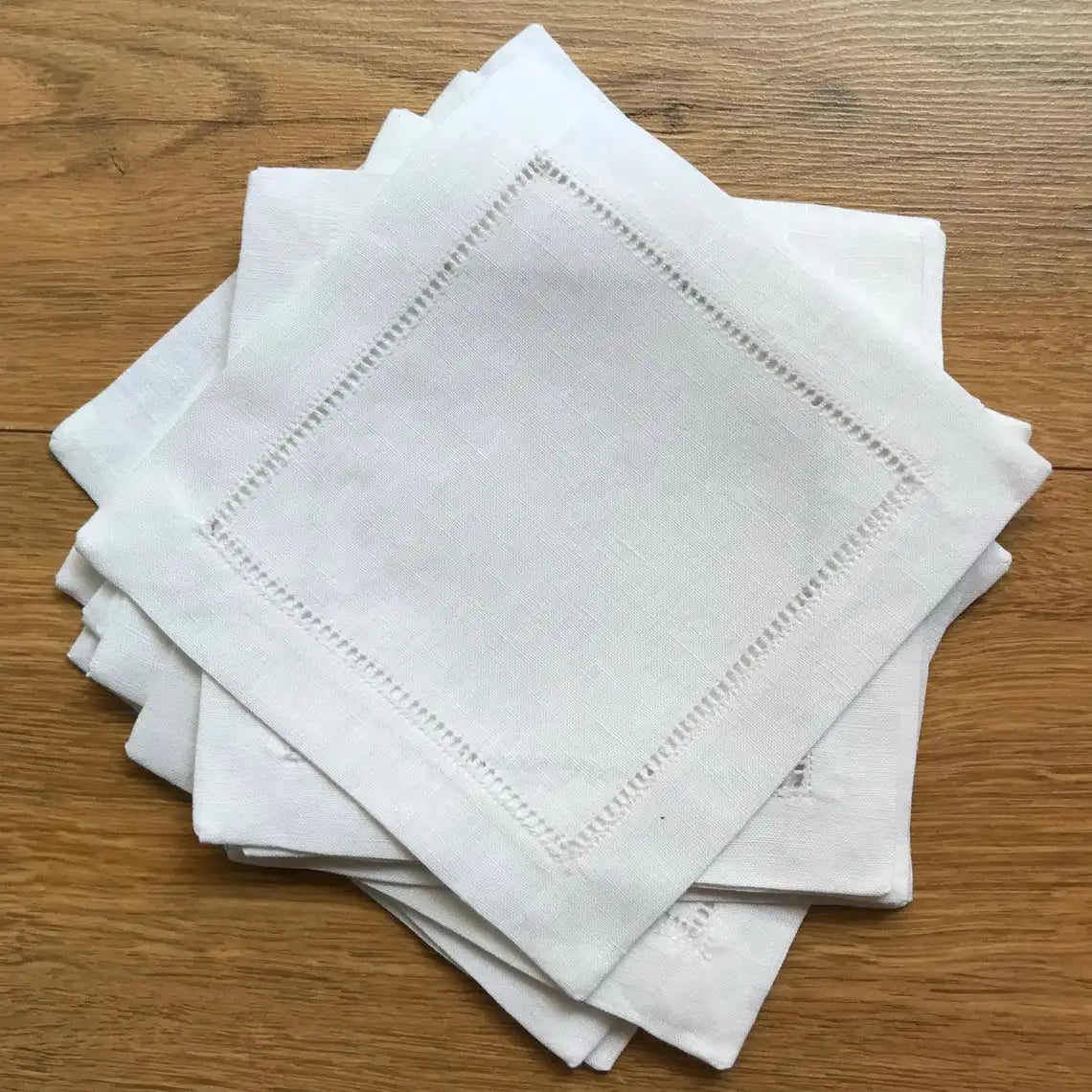 Pile de serviettes blanches en lin avec un design élégant de motifs perlés brodés autour des bords, reposant sur une table en bois.