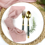 Une serviette de table rose, une fourchette et une cuillère dorées posées sur un plateau en osier, entourées de verdure fraîche.