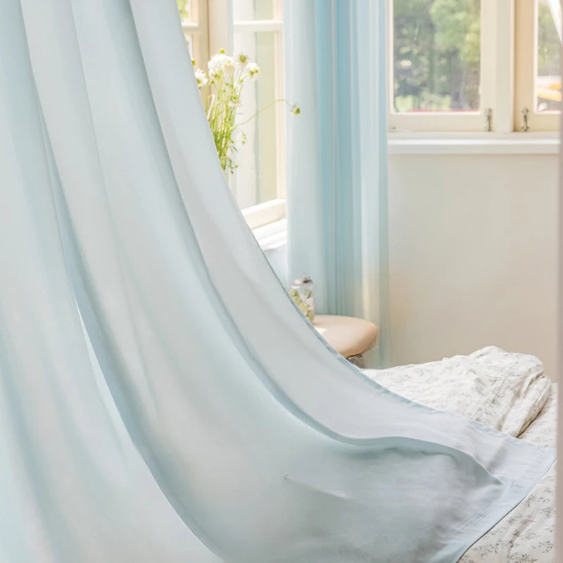 Voilage bleu translucide flottant délicatement devant une fenêtre ouverte, apportant une lumière douce à une chambre lumineuse.