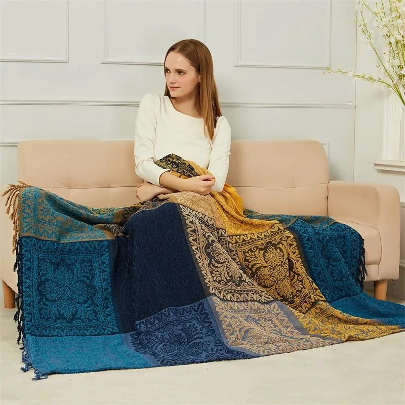 La jeune femme est assise sur un canapé beige et est enveloppée dans une couverture colorée ornée de motifs ethniques, créant une atmosphère cocooning et apaisante.