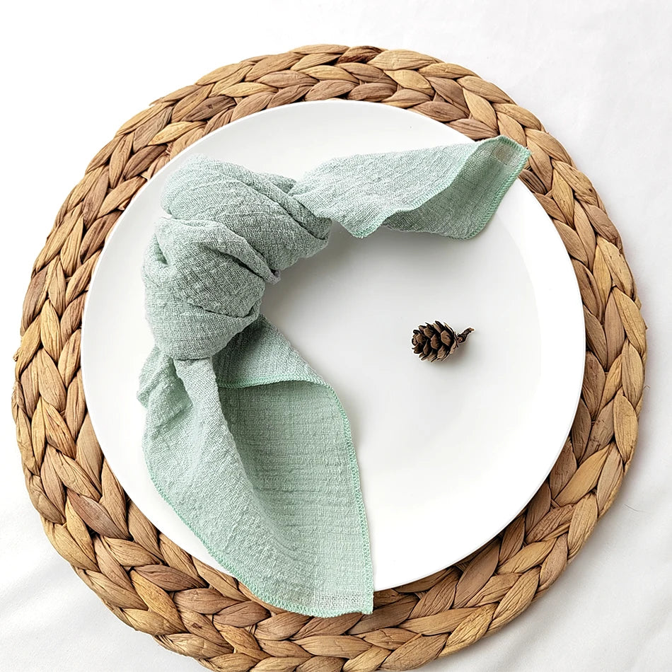 Un plat circulaire en osier tressé accueille une serviette de couleur verte pastel et un petit cône de pin, créant une atmosphère chaleureuse et naturelle.