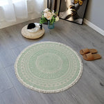 Un tapis rond vert pâle décoré de motifs complexes, posé sur un sol en bois gris, avec un panier en forme de chat et des sandales à côté.