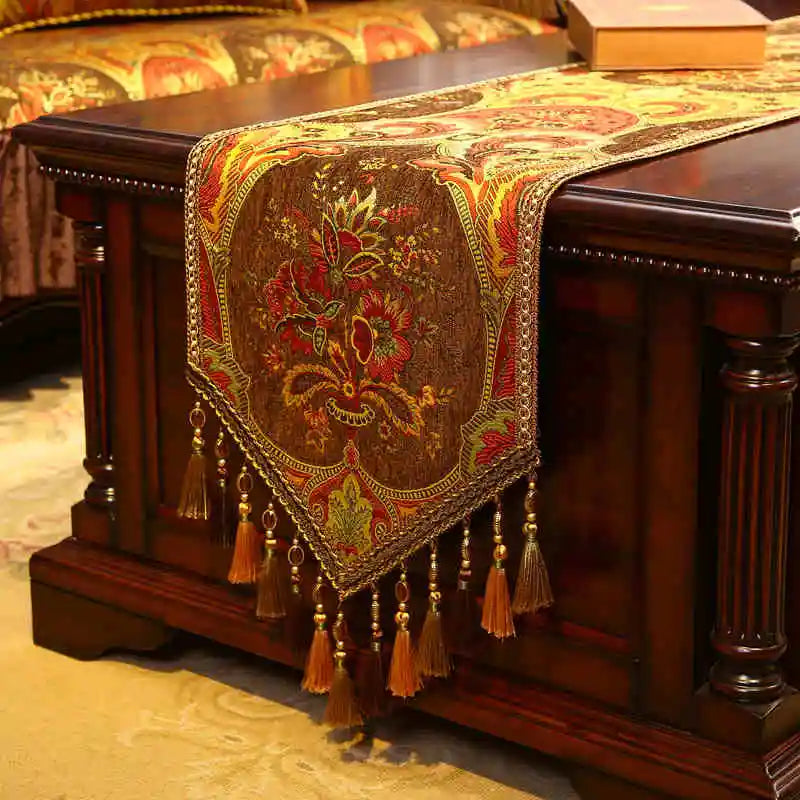 Élégant chemin de table en tissu brocart doré et rouge, orné de motifs floraux, de franges et de glands décoratifs, posé sur un meuble en bois foncé.