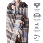 Une femme enveloppée dans une couverture à carreaux grise et beige qui peut être utilisée comme un châle pour les épaules, une couverture pour le canapé ou le lit, ainsi qu'un accessoire de voyage.