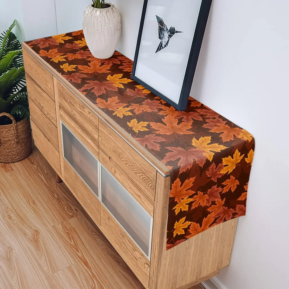 Cette image montre un meuble en bois avec un plateau décoré de motifs de feuilles d'automne colorées, dans des tons de brun, d'orange et de jaune. Le meuble est placé dans un intérieur décoré avec d'autres éléments naturels comme un vase et des plantes.