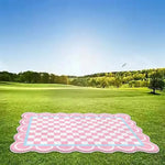 Un tapis de pique-nique rose et blanc posé sur un pré verdoyant, entouré d'arbres et d'un ciel bleu, créant une scène idyllique et sereine.
