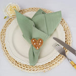 Un élégant arrangement de table avec une serviette de table verte pliée et décorée d'un cœur en rotin, accompagné d'une fourchette et d'un couteau, sur un napperon tressé beige.