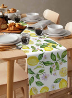 La table de petit-déjeuner est garnie de produits de boulangerie, de vaisselle blanche et de verres, le tout posés sur une nappe imprimée de motifs végétaux aux couleurs fraîches et ensoleillées.