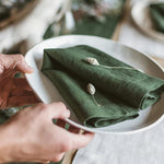 Sur une assiette blanche, une serviette en tissu vert est pliée avec soin, avec quelques petites fleurs blanches posées dessus, mettant en valeur la texture et la couleur de la serviette.