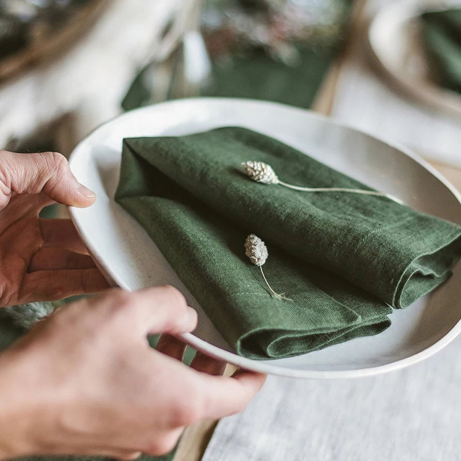 Sur une assiette blanche, une serviette en tissu vert est pliée avec soin, avec quelques petites fleurs blanches posées dessus, mettant en valeur la texture et la couleur de la serviette.