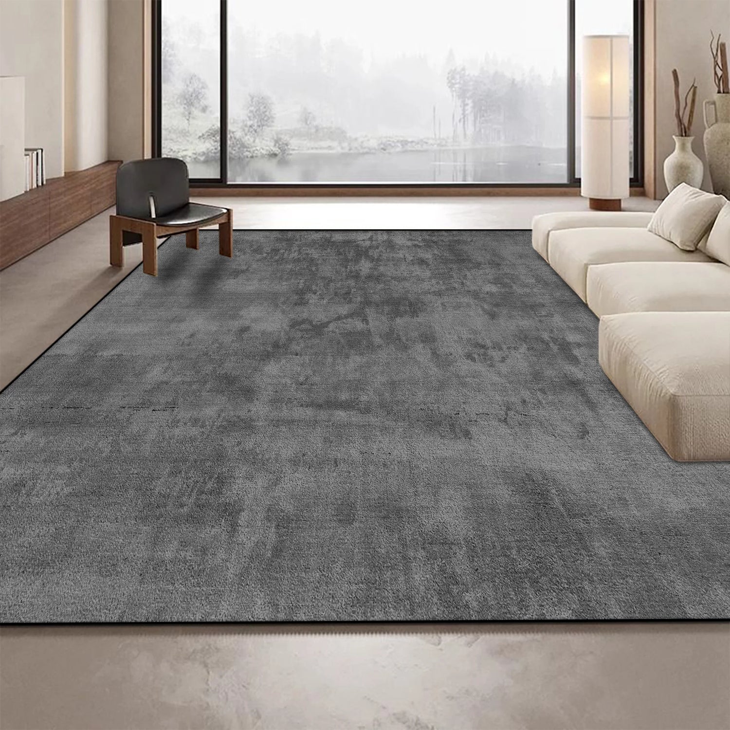Un tapis gris foncé recouvre le sol d'un salon spacieux avec de grandes fenêtres offrant une vue sur un paysage forestier enneigé.
