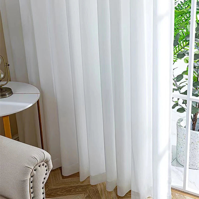 Voilage blanc posé dans un salon avec fauteuil capitonné beige, petite table ronde blanche et parquet en chevrons près d’une baie vitrée.