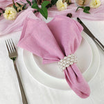 Une élégante serviette rose ornée d'une jolie broche de perles sur une assiette blanche avec couverts et fleurs d'automne, suggérant un élégant couvert de table.