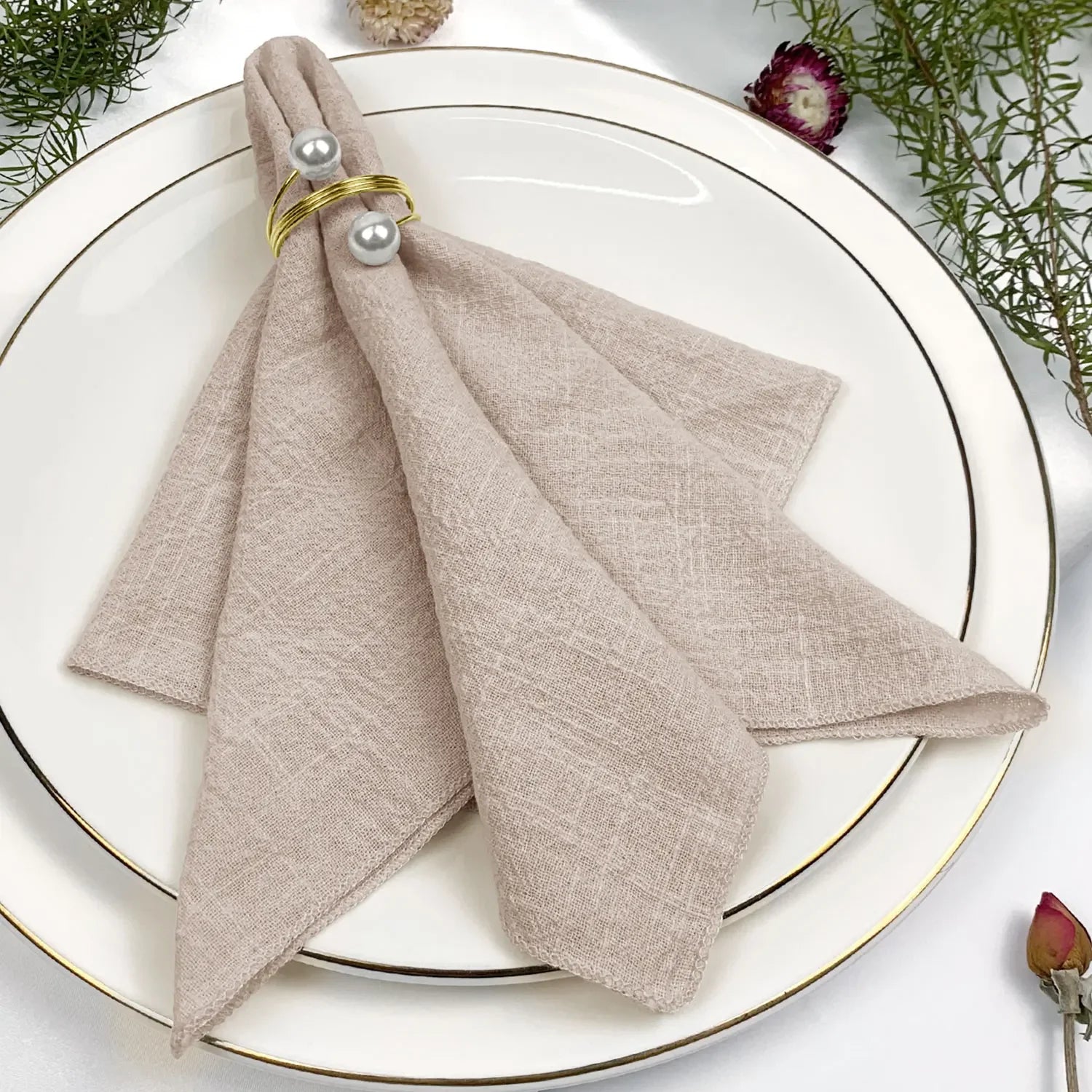 Une élégante serviette de table beige accompagnée d'anneaux dorés et d'accents végétaux, créant une décoration de table raffinée.