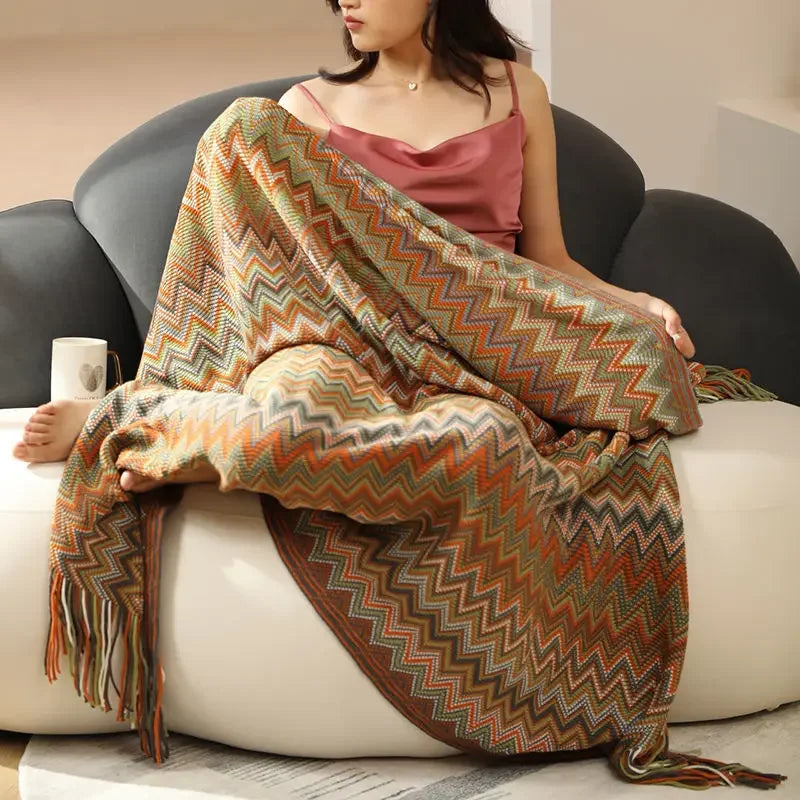 Cette image montre une jeune femme assise sur un canapé, portant une robe rose et enveloppée dans une couverture à motif géométrique en couleurs chaudes. La couverture est ornée de franges et semble avoir un aspect texturé et bohème.