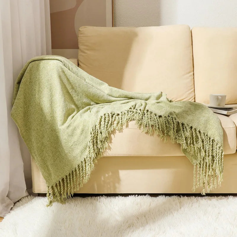 Couverture en tricot moelleux de couleur vert pistache avec des franges décoratives, placée sur un canapé crème dans un intérieur douillet.