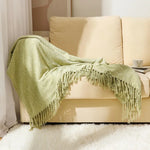 Couverture en tricot moelleux de couleur vert pistache avec des franges décoratives, placée sur un canapé crème dans un intérieur douillet.