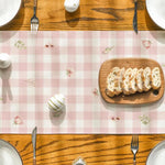 Sur cette image, on peut voir une table en bois avec un élégant napperon à carreaux roses et blancs, sur lequel sont posés des couverts, une assiette et une tranche de pain rustique sur une planche de bois.
