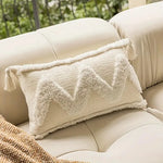 Coussin décoratif avec motifs triangulaires et franges texturées, posé sur un canapé beige dans un environnement naturel.