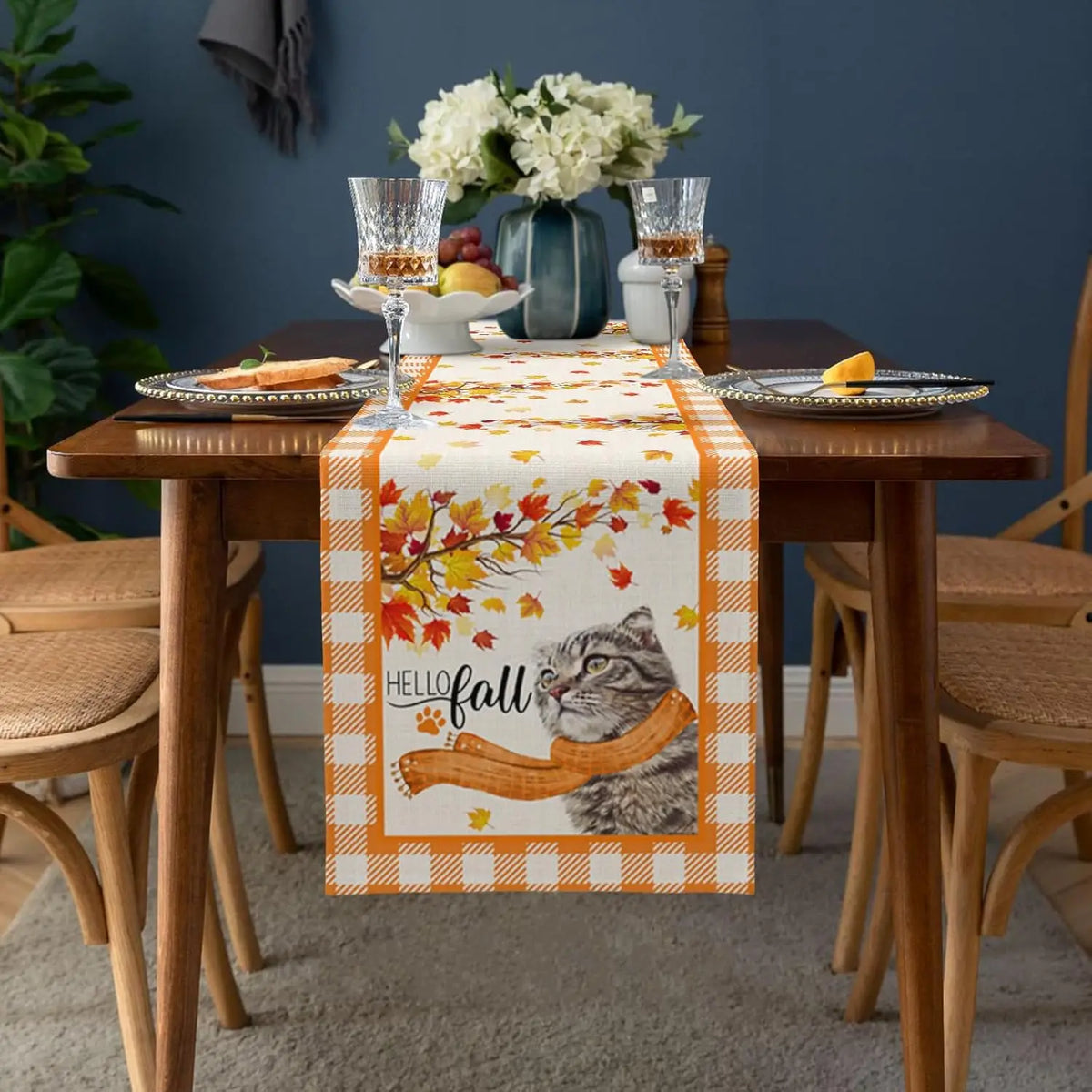 Une belle table d'automne avec un centre-de-table fleuri, des verres de cristal, des fruits et un joli chemin de table aux motifs de feuilles d'automne et d'un chat.