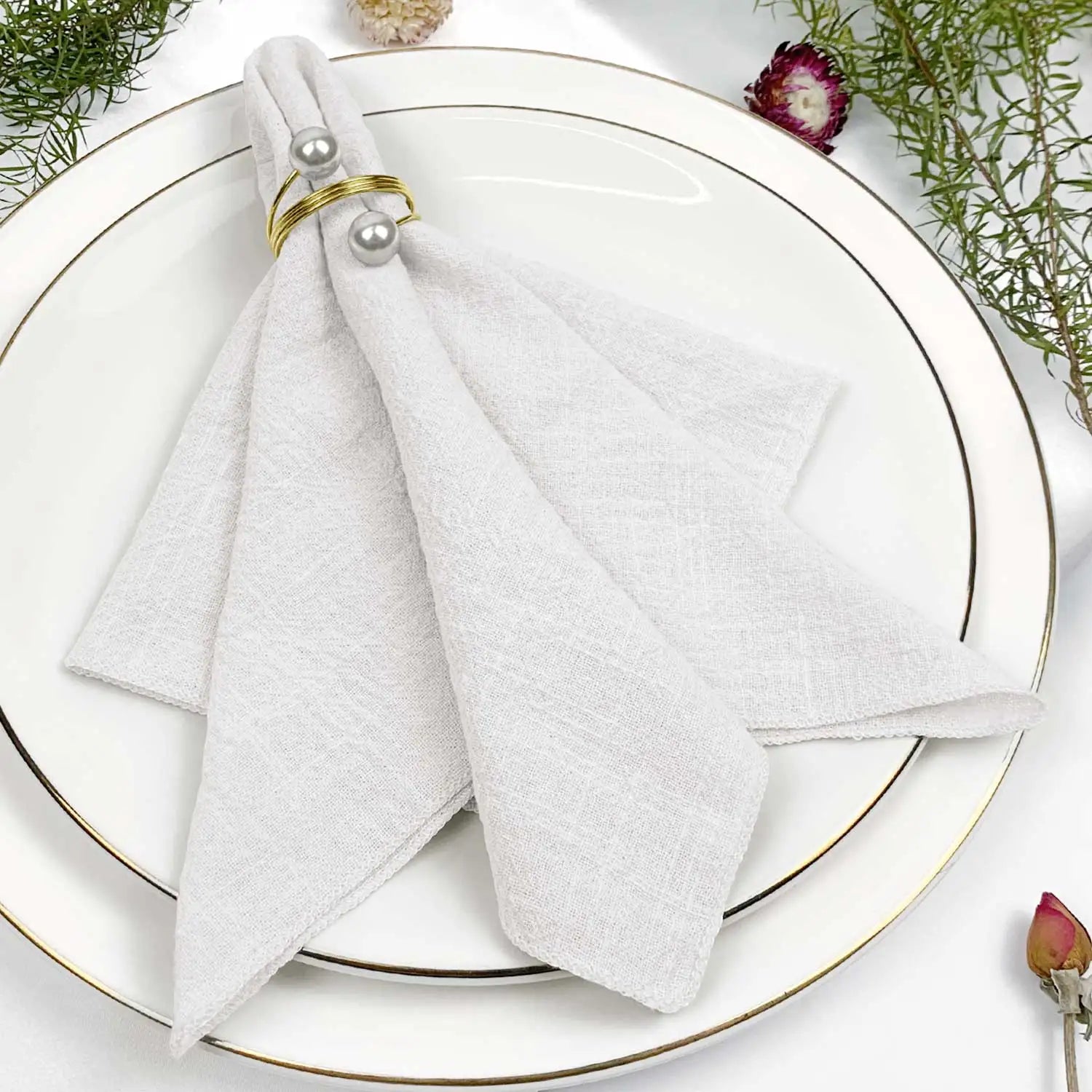 Serviette blanche élégamment disposée sur une assiette blanche avec des anneaux dorés et des fleurs décoratives.