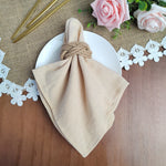 Serviette de table beige nouée avec une corde, placée sur une nappe en jute, entourée de roses roses, de feuilles vertes et de petites fleurs blanches.