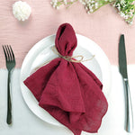 Une serviette de table de couleur bordeaux, élégamment pliée et attachée avec une ficelle, sur une assiette blanche, entourée de fleurs blanches sur un fond rose pâle.