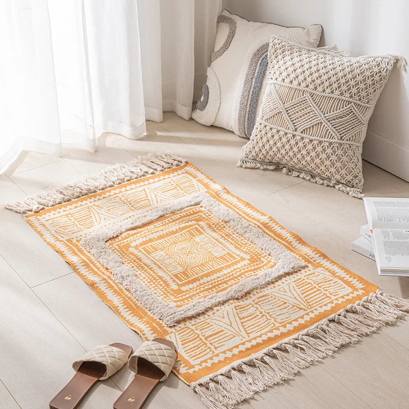 Un tapis décoratif de style bohème avec des motifs géométriques et une frange en lin, posé sur un sol clair à côté de coussins décoratifs.