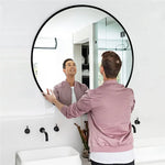 Un homme souriant regarde son reflet dans un miroir rond noir dans une salle de bain lumineuse et épurée.