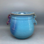 Un pot en céramique bleu clair avec des ornements dorés et des poignées violettes, offrant un bel objet décoratif de style vintage.