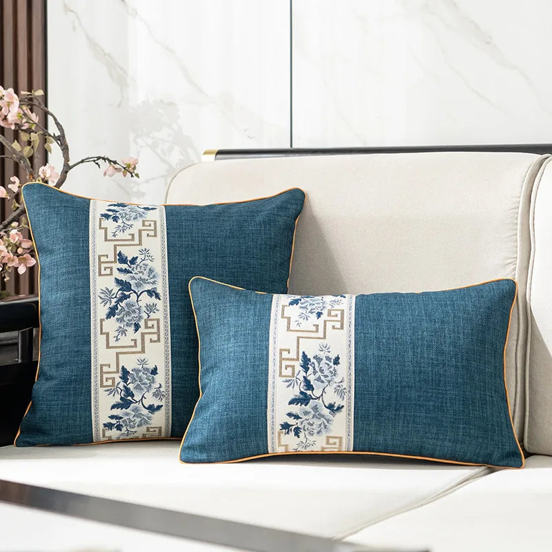 Deux coussins décoratifs aux motifs floraux bleus et blancs, ornés de bordures ornées, placés sur un canapé blanc dans un intérieur lumineux.