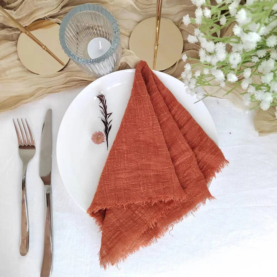 Sur une assiette blanche, une serviette de table orange foncé aux bords effilochés est joliment disposée, accompagnée de couverts en métal et d'une composition florale avec des fleurs blanches.