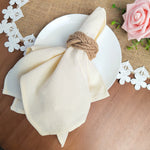 Sur un fond de jute brun, on voit une assiette blanche avec un linge blanc plié et noué avec de la corde de jute, entouré de fleurs blanches en papier et d'une rose rose.