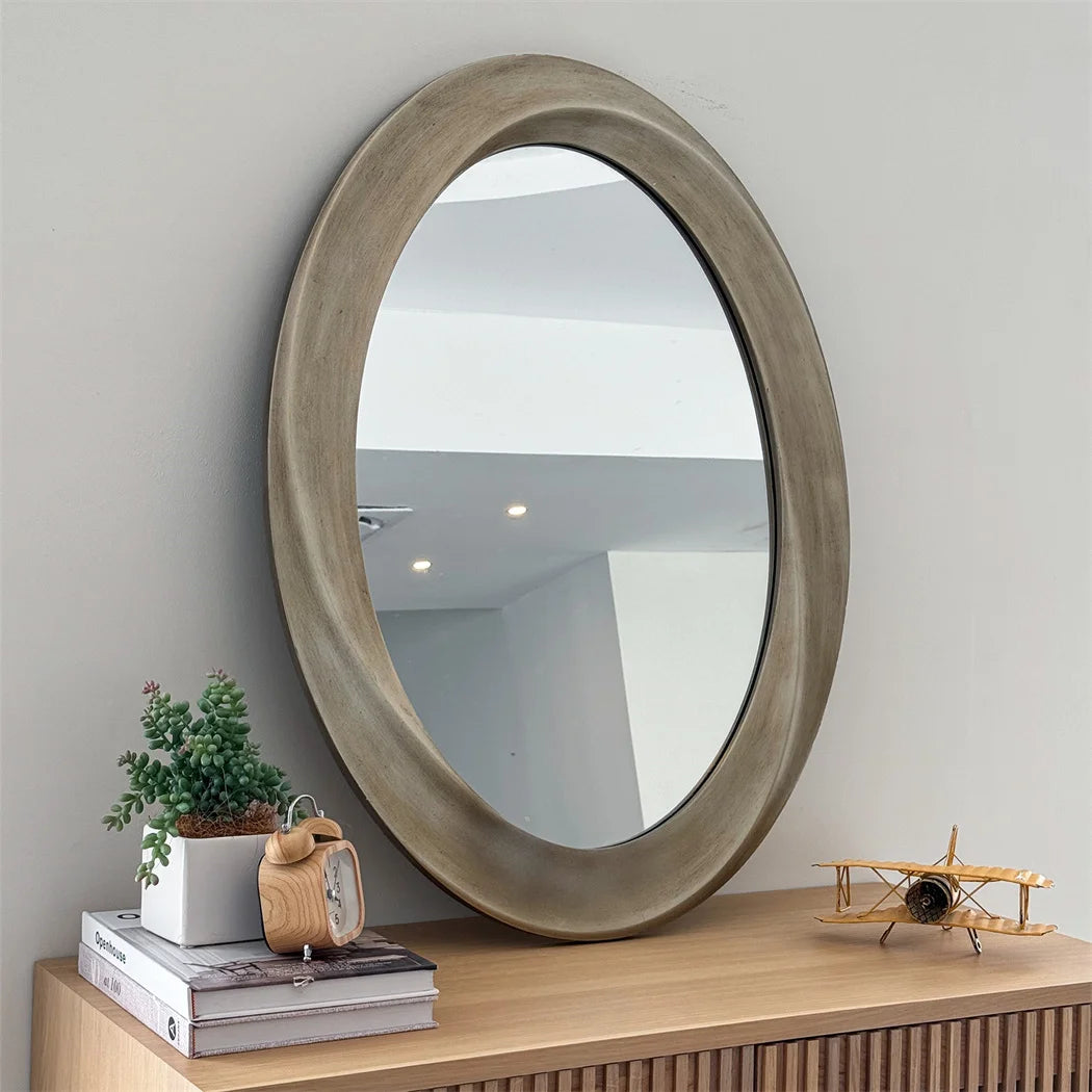 Un miroir ovale à cadre en bois gris posé sur un meuble en bois clair, accompagné de décorations telles qu'une petite plante en pot et un avion en bois.