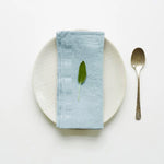 Une serviette de table bleu clair, décorée d'une feuille verte, est posée sur une assiette blanche, accompagnée d'une cuillère en métal.