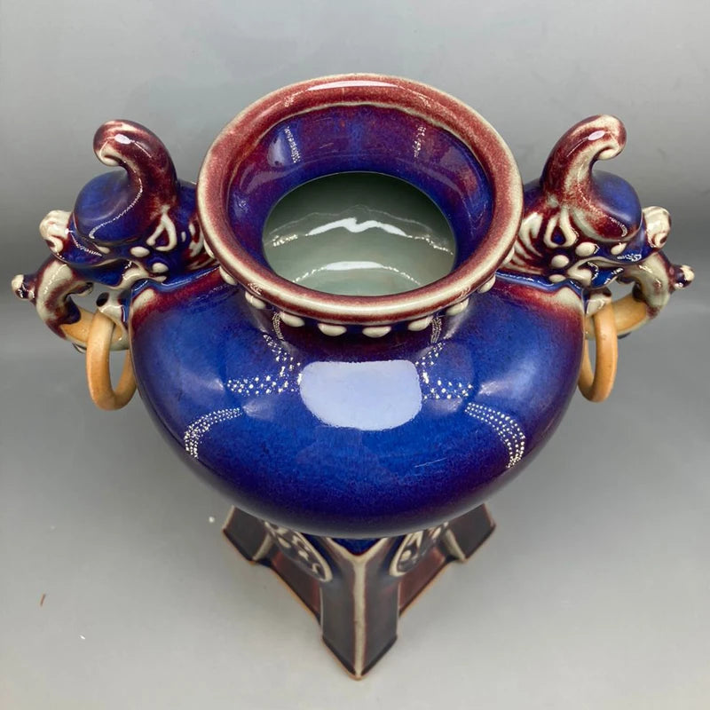 Un bol en céramique bleu foncé décoré de motifs dorés, avec deux appendices latéraux en forme de têtes animales, posé sur un support en métal.