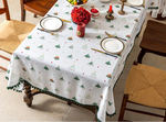 Sur cette image, une décoration de table de fête est visible. La table est recouverte d'une nappe blanche avec des motifs d'arbres de Noël verts et rouges. Des assiettes, des couverts et un arrangement floral composé de fleurs rouges et de baies ornent la table.