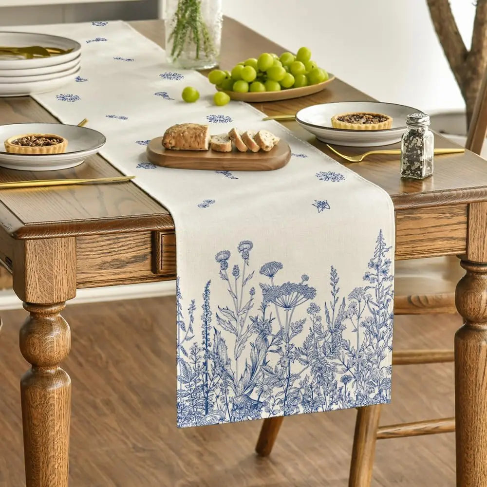 Cette image présente une nappe de table blanche avec un imprimé floral bleu, sur laquelle se trouvent des assiettes, des verres, un plat de raisin vert et des tartes dans un cadre rustique en bois.
