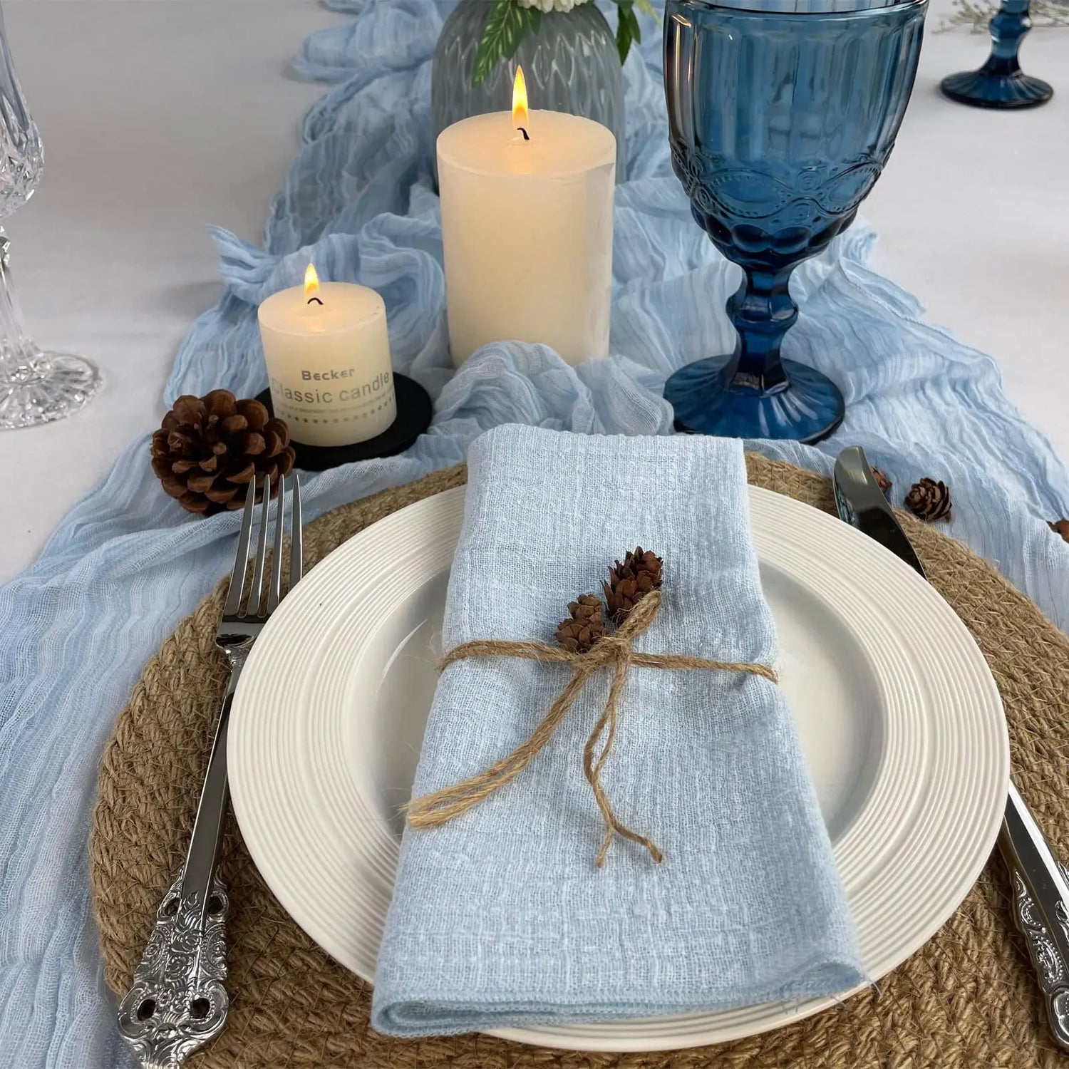 Cette image représente une table de fête élégante avec des bougies allumées, un verre bleu, des cônes de pin et une serviette grise nouée avec une ficelle. Cette mise en scène chaleureuse et naturelle crée une ambiance conviviale et raffinée pour un repas ou une réception.